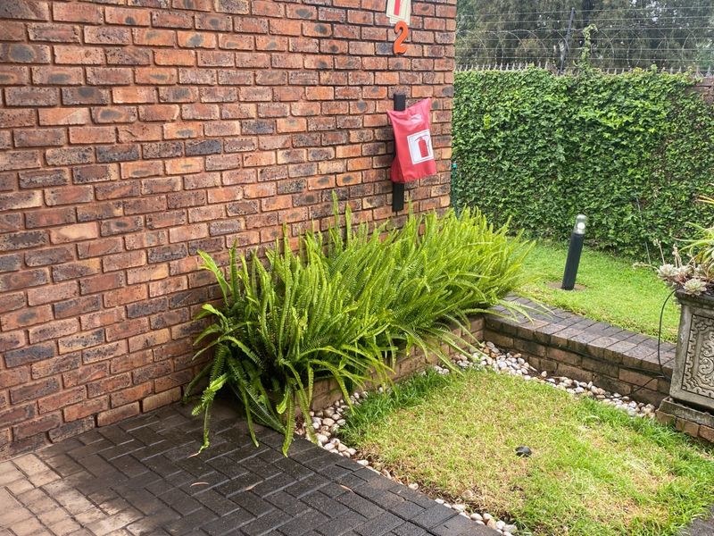 A view of the apartment exterior featuring a face brick wall, paved walkway, and landscaped garden area with an electric fence boundary.