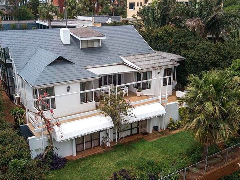 Aerial view of a residential property featuring a multi-level roof, rear balcony, paved patio areas, and established garden surroundings.