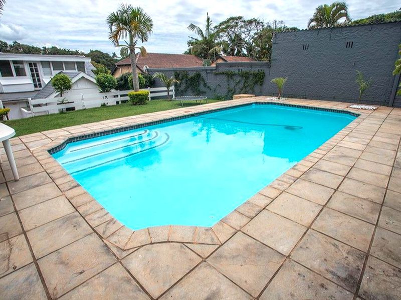 A well-maintained outdoor swimming pool area featuring a paved deck, surrounding lawn, and a secure boundary wall.
