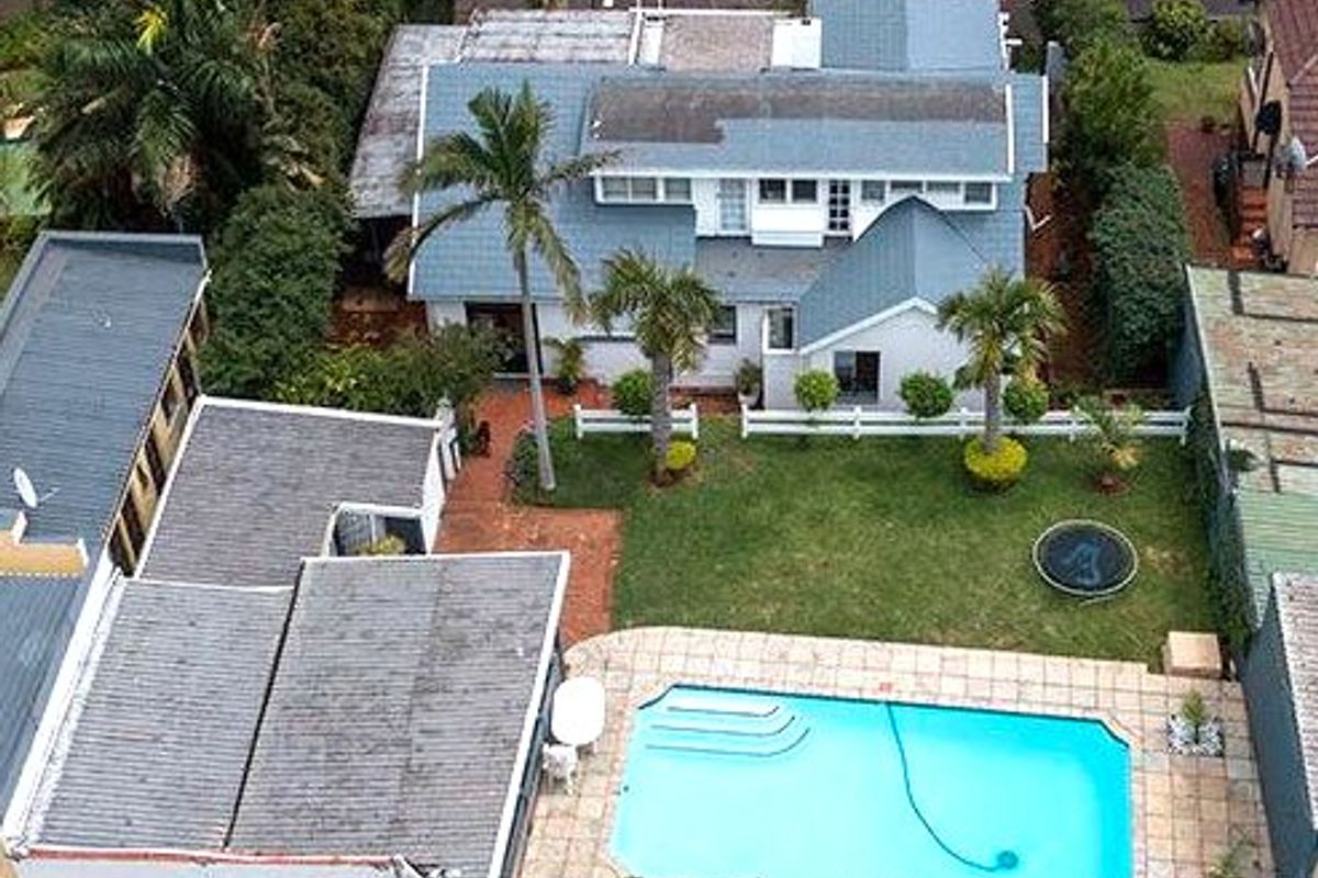 Aerial view of a residential property featuring a swimming pool, landscaped garden, detached garage, and a multi-level house with a pitched roof.
