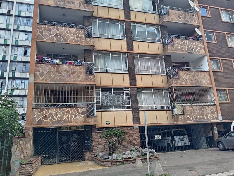Front elevation of a multi-story apartment building featuring stone cladding, balconies with security bars, and a ground-level gated entrance.