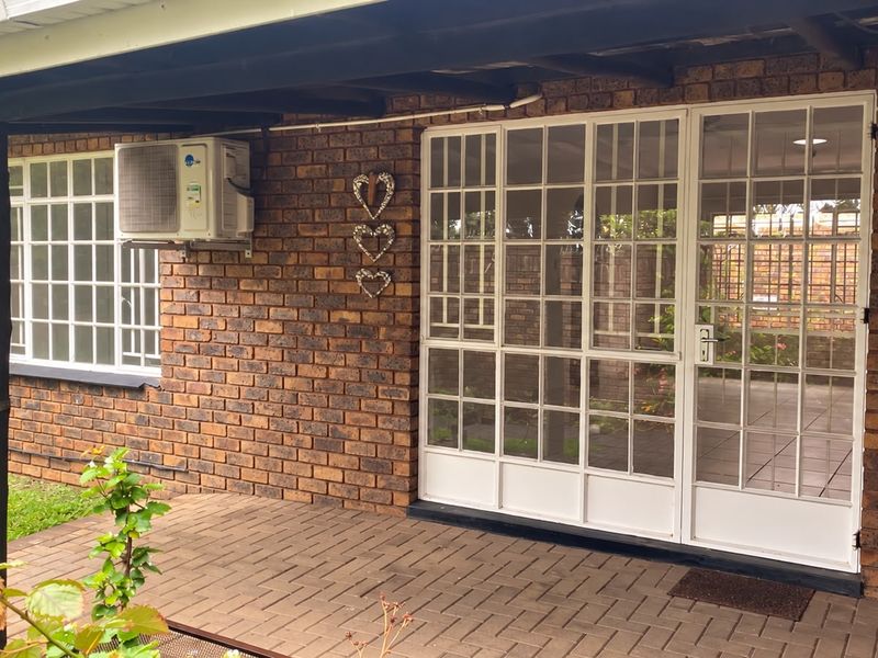 A covered brick patio area featuring paved flooring, a wall-mounted air conditioning unit, and secure grid-pattern glass doors leading into the apartment.