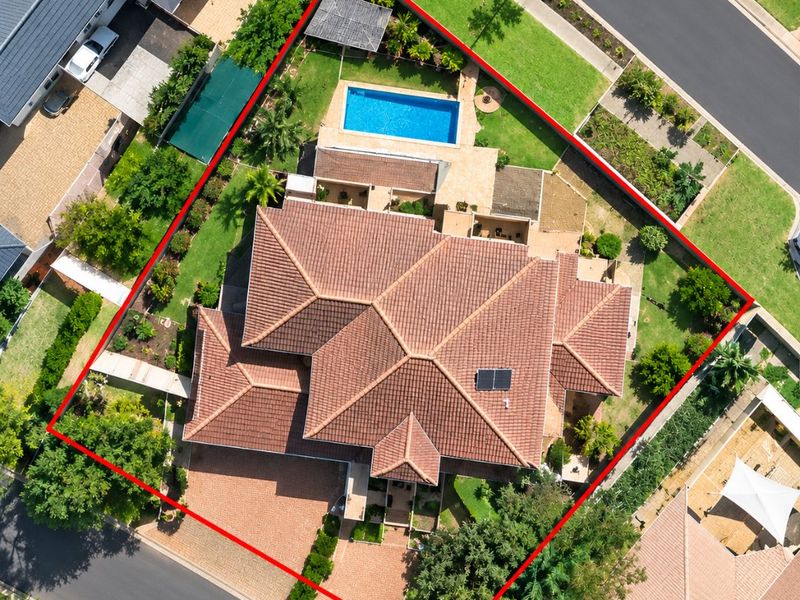 Aerial view of a residential property featuring a tiled roof, swimming pool, solar panel, paved driveway, and established garden within a fenced perimeter.
