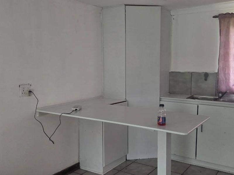 A basic kitchen area featuring built-in white cabinetry, a stainless steel sink, tiled backsplash, and a breakfast counter, requiring cosmetic updates.