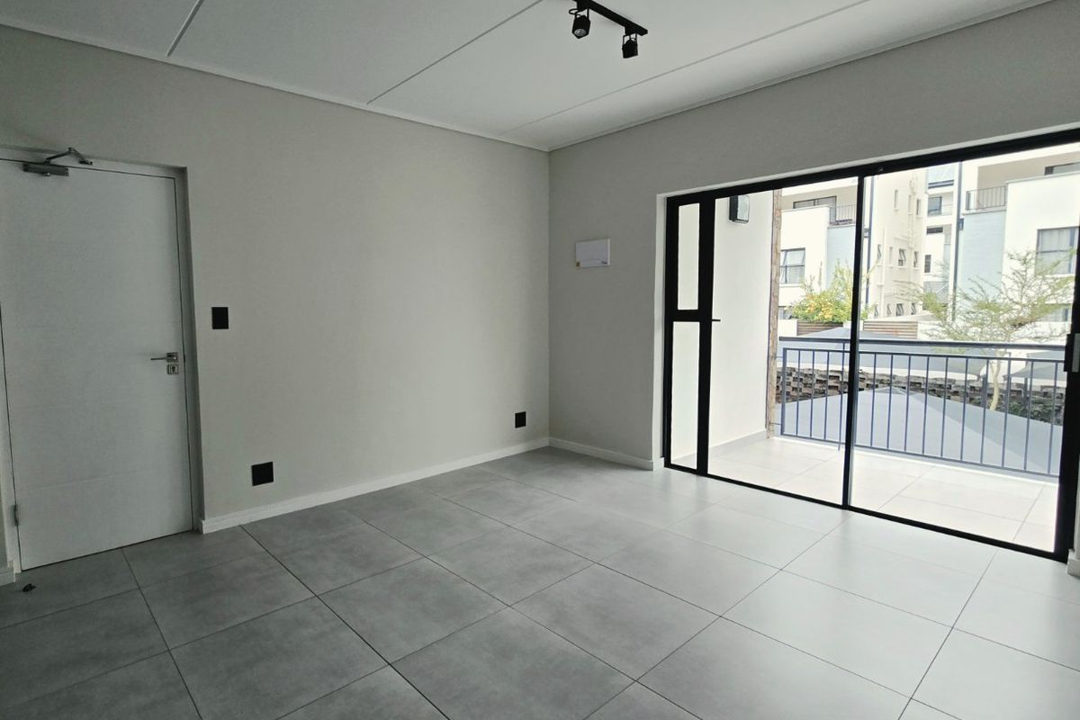 A bright, modern, and empty living area featuring large format grey floor tiles, track lighting, and a sliding glass door leading to a balcony.