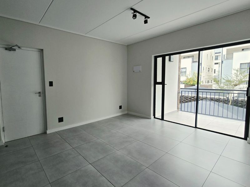 A bright, modern, and empty living area featuring large format grey floor tiles, track lighting, and a sliding glass door leading to a balcony.