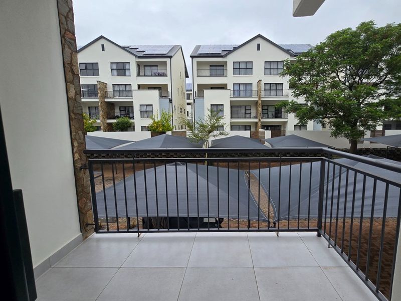 A clean, tiled balcony featuring a metal balustrade and stone-clad pillar, overlooking a residential complex with shaded parking bays.
