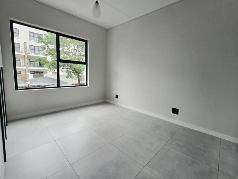 A bright, empty bedroom featuring modern large-format floor tiles, neutral walls, and a large window providing natural light.