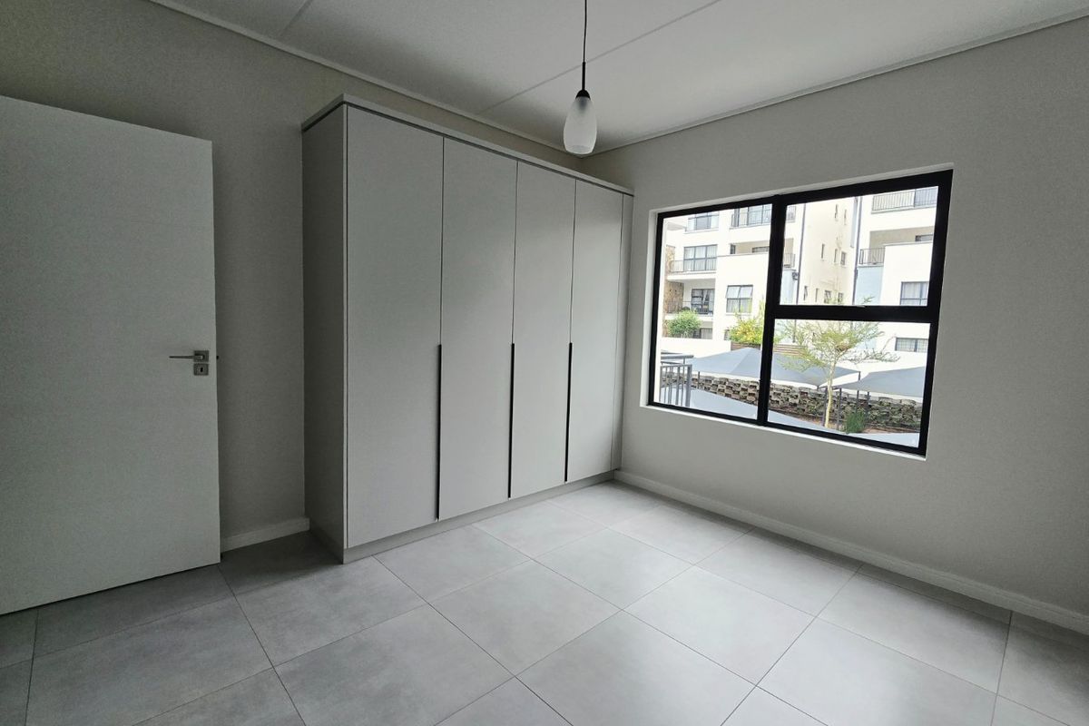A bright, modern bedroom featuring neutral tiled flooring, a sleek built-in wardrobe, and a large window with contemporary black aluminium frames.