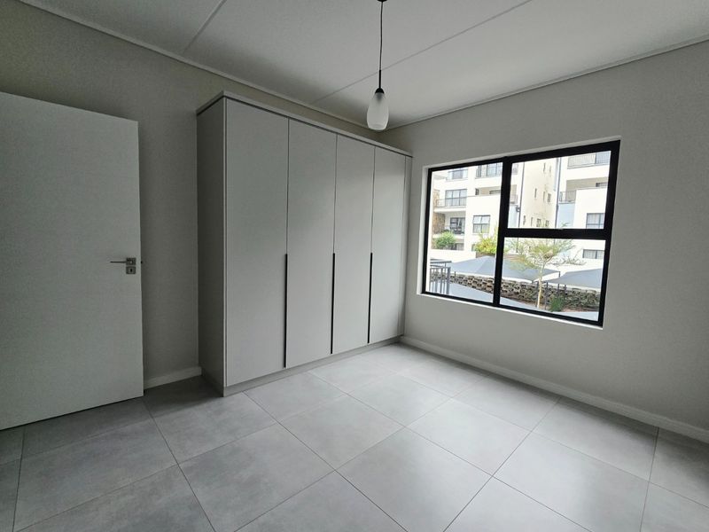 A bright, modern bedroom featuring neutral tiled flooring, a sleek built-in wardrobe, and a large window with contemporary black aluminium frames.