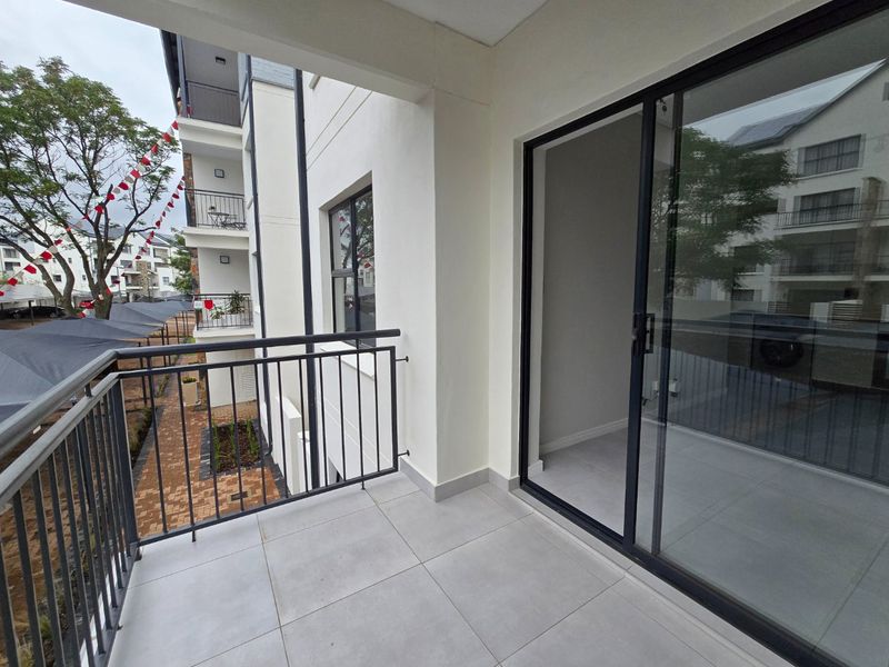 A clean, modern balcony featuring light-toned floor tiles, a secure metal balustrade, and a large sliding glass door providing access to the interior.