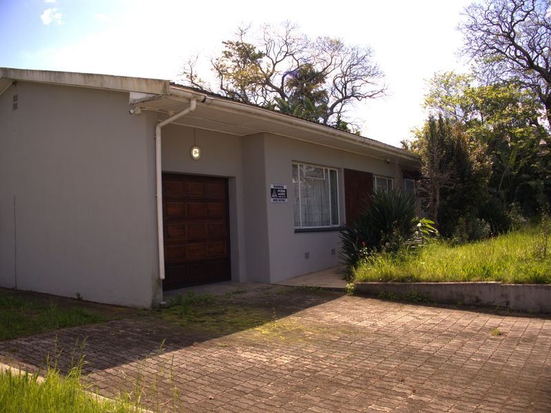 Front exterior view of a single-story house featuring a paved driveway, integrated garage, and established garden area requiring maintenance.