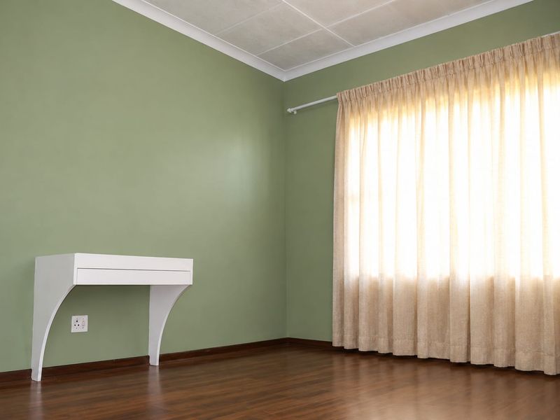 A bright, empty bedroom featuring green walls, dark laminate flooring, and a large window with installed curtain rails.