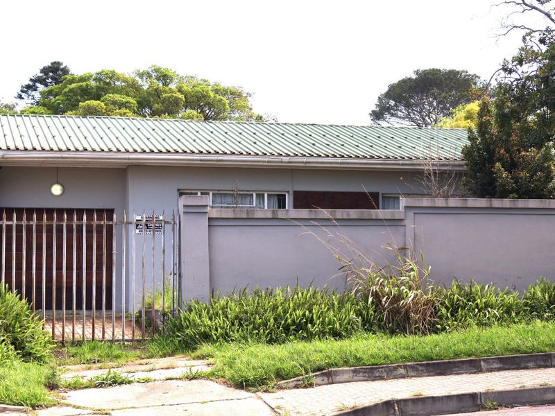 Front elevation of a single-story house featuring a metal roof, perimeter wall, and gated driveway access with visible signs of exterior maintenance required.