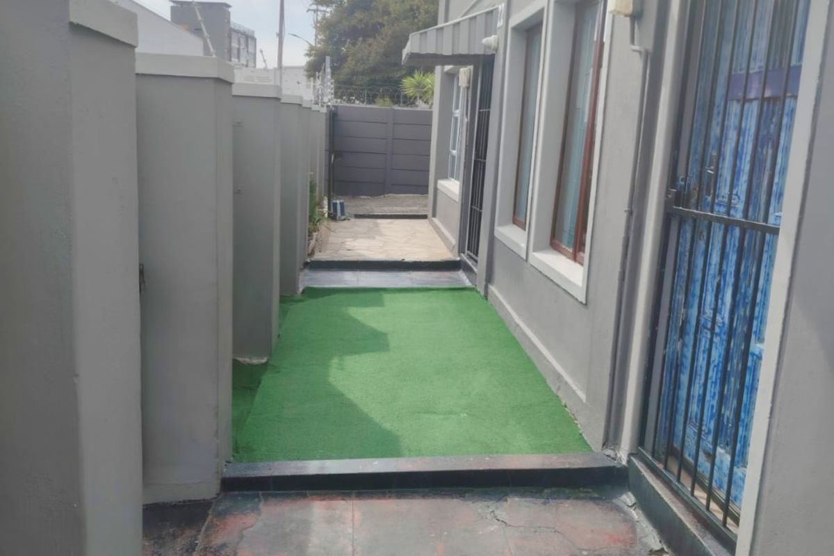 A side passage featuring a tiled walkway, artificial grass section, security gate, and protective awnings along the building exterior.