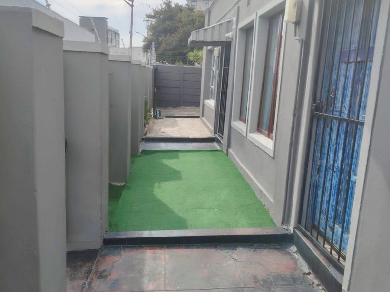 A side passage featuring a tiled walkway, artificial grass section, security gate, and protective awnings along the building exterior.