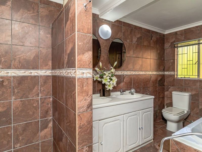 A functional bathroom featuring a shower, double vanity, toilet, and bathtub with full-height brown wall tiling and security bars on the window.