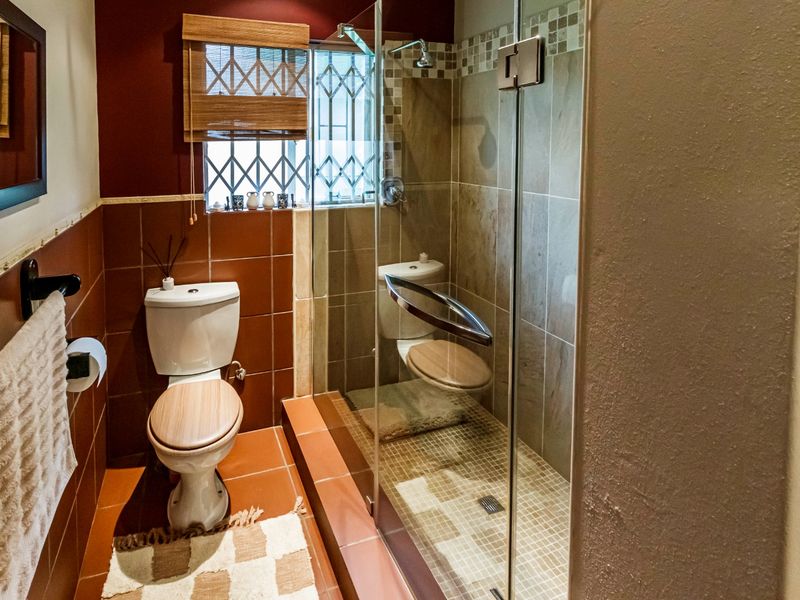 A compact bathroom featuring a glass-enclosed shower, tiled walls, and a standard toilet, with a window fitted with a security gate.