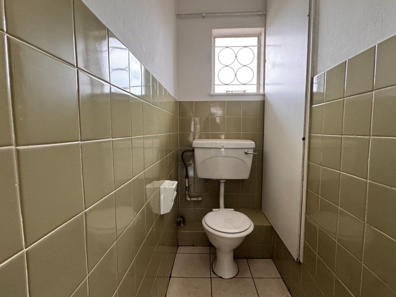 A compact, dated bathroom featuring olive-toned ceramic wall tiles, tiled flooring, and a standard toilet with security bars on the window.