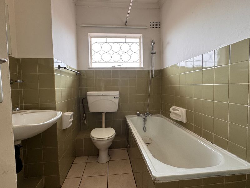 A dated bathroom featuring olive green wall tiles, a standard bathtub, pedestal basin, and toilet, with a security-grille window for ventilation.