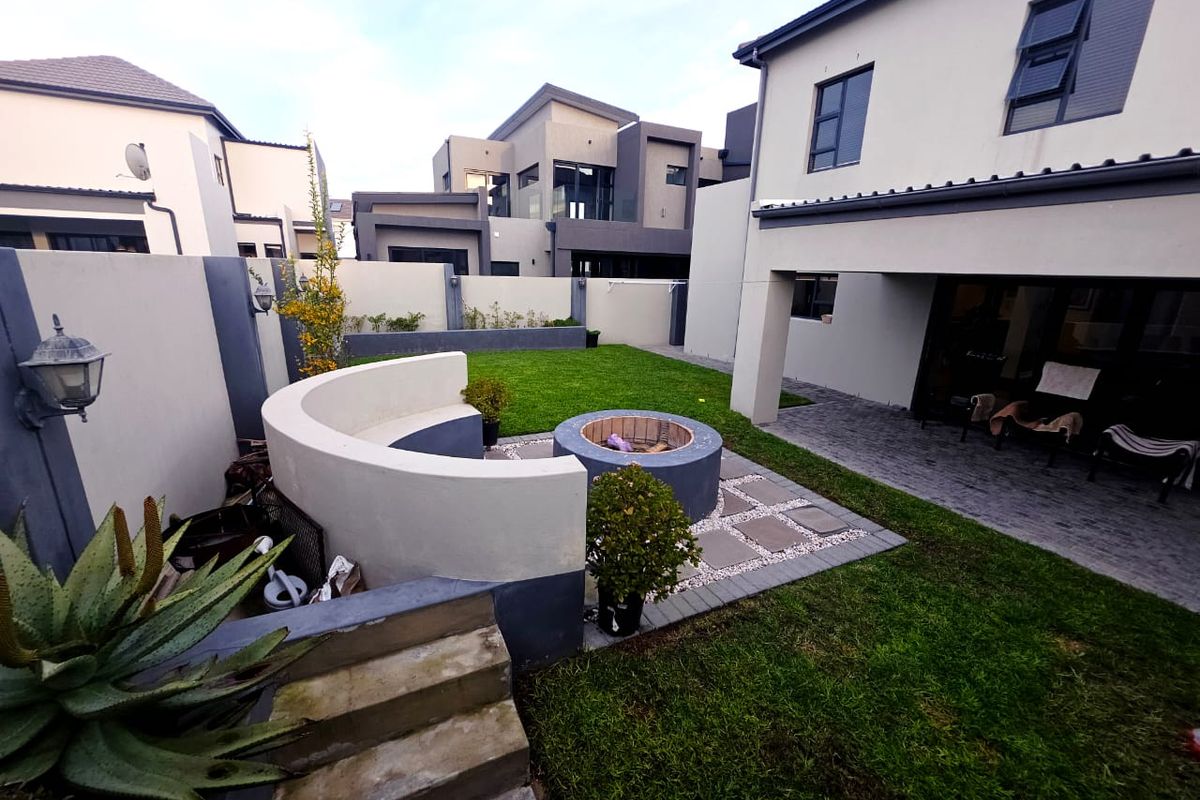 A well-maintained rear garden featuring a paved patio, a built-in circular fire pit with a curved concrete bench, and a manicured lawn.
