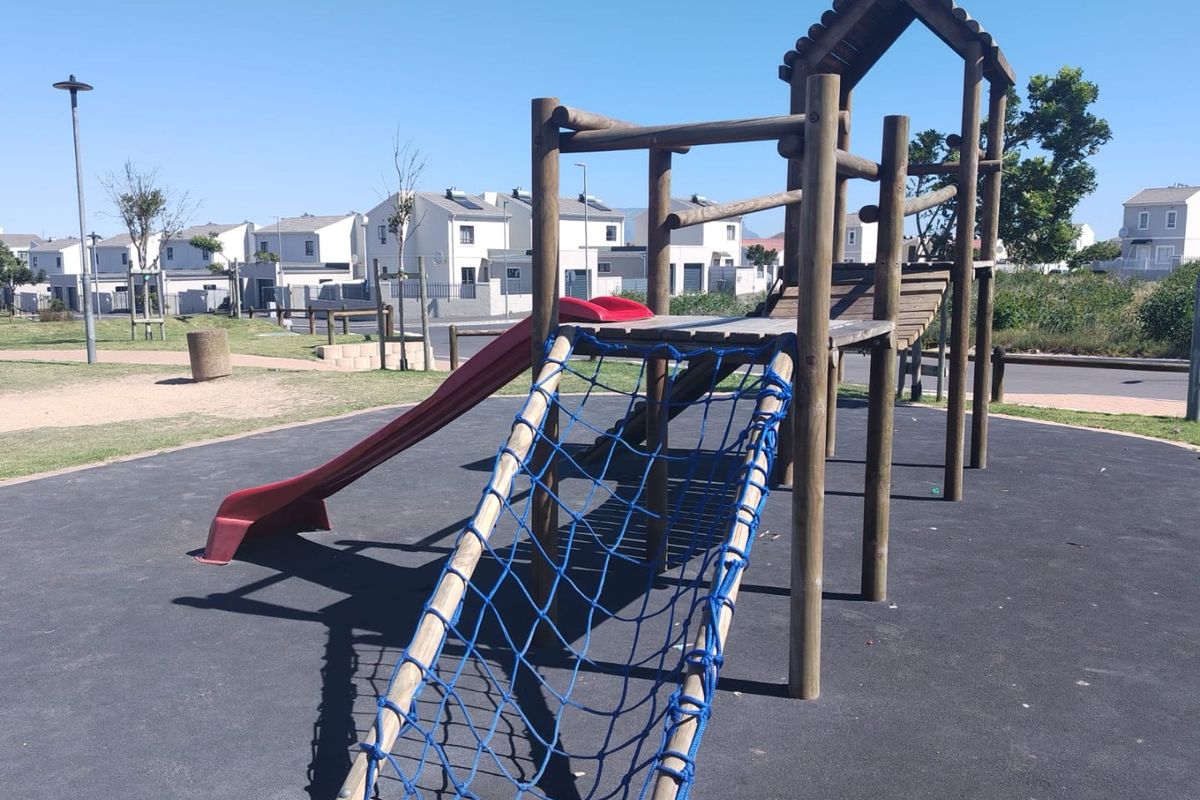 A communal outdoor playground area featuring a wooden climbing structure with a slide and rope net, set on a rubberized safety surface.