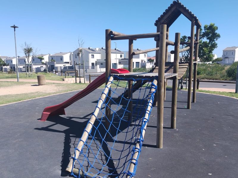 A communal outdoor playground area featuring a wooden climbing structure with a slide and rope net, set on a rubberized safety surface.