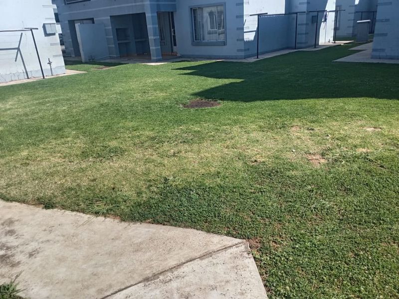 A rear exterior view of the property featuring a shared lawn area, concrete pathways, and a built-in outdoor braai facility.