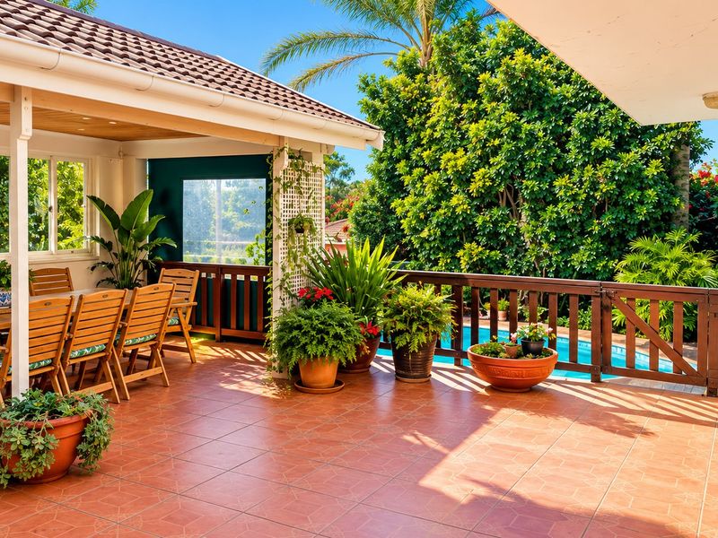 A spacious covered patio featuring tiled flooring and a wooden balustrade, overlooking a private swimming pool and lush garden area.