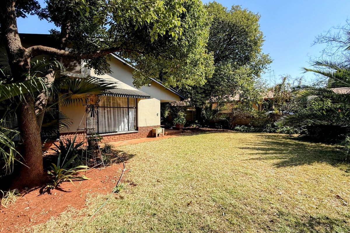 A spacious rear garden featuring a well-maintained lawn, mature trees, and a paved patio area adjacent to the house facade.