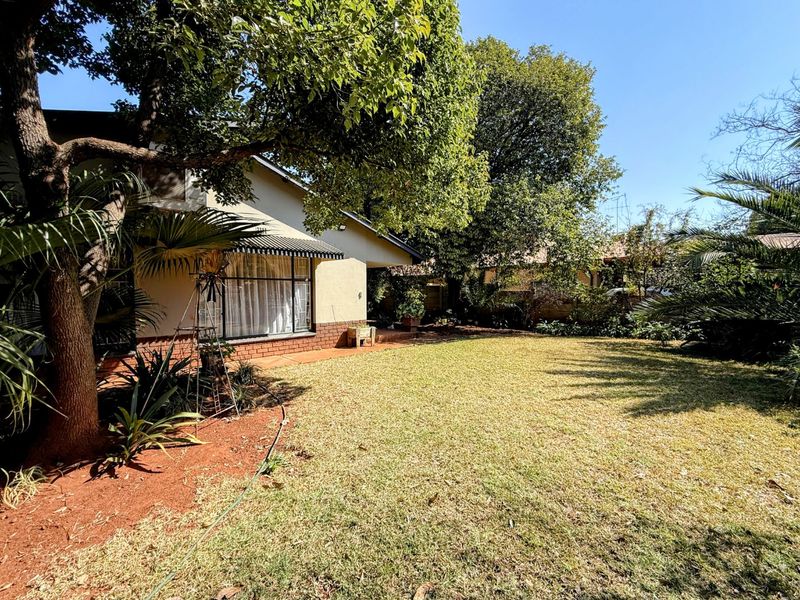 A spacious rear garden featuring a well-maintained lawn, mature trees, and a paved patio area adjacent to the house facade.
