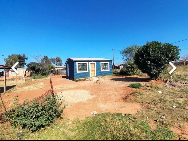 A detached single-story dwelling featuring a painted brick exterior, corrugated metal roof, and aluminium window frames, situated on an unpaved plot.
