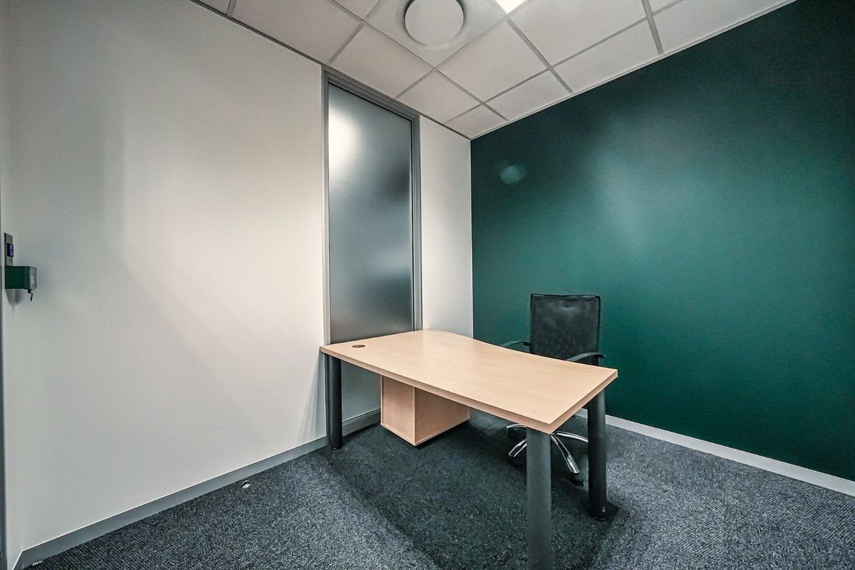 A compact, modern office space featuring a dark green accent wall, suspended ceiling, and frosted glass partition, providing a professional work environment.