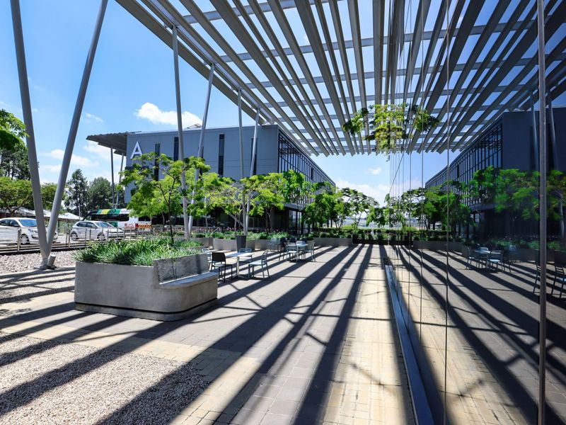 A modern commercial outdoor patio area featuring a large louvred pergola, paved surfaces, and integrated concrete planters adjacent to a glass-fronted office building.