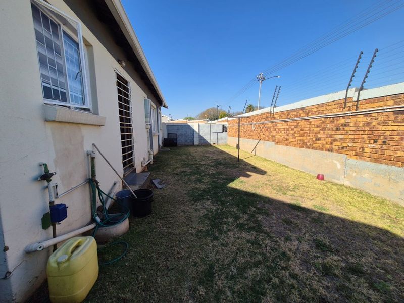 A rear exterior view of the property featuring a lawn area, brick boundary wall with electric fencing, and security features on the building facade.