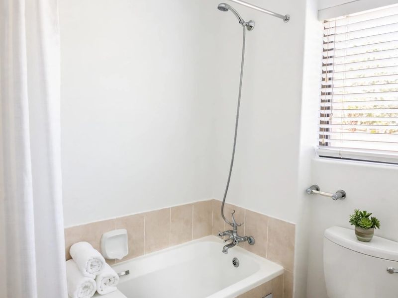 A clean, functional bathroom featuring a bathtub with a handheld shower mixer, tiled splashback, and a window with venetian blinds.