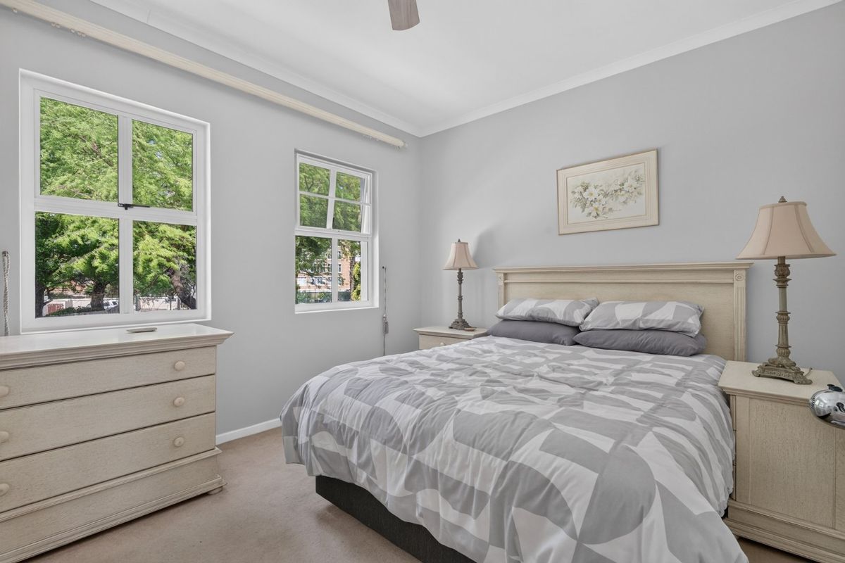 A well-maintained bedroom featuring neutral wall tones, carpeted flooring, and dual casement windows providing natural light and a pleasant outlook.