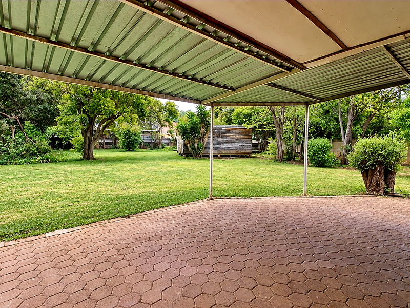 A covered outdoor patio area featuring paved flooring and a metal roof structure, overlooking a spacious garden with mature trees.