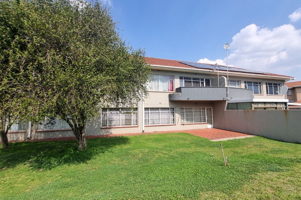 Rear view of the apartment building featuring a private garden, paved patio area, and an upper-level balcony with solar panels installed on the roof.