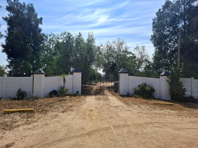 A gated entrance featuring white perimeter walls with concrete pillars and a metal gate, leading onto a dirt driveway.