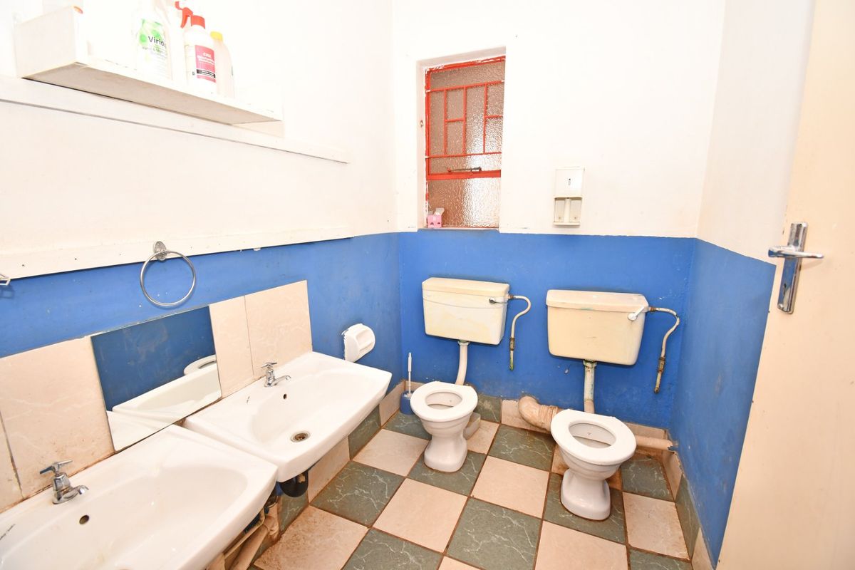 A functional commercial bathroom featuring dual toilets and washbasins with tiled flooring and security-barred window, requiring cosmetic updates and maintenance.