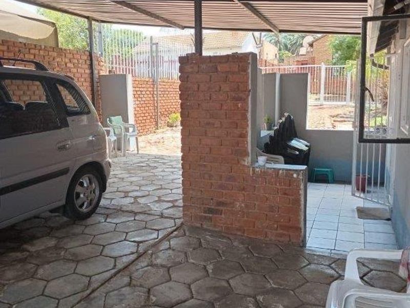 A covered outdoor parking area featuring a corrugated metal roof, brick partition wall, and paved flooring, providing functional shelter for a vehicle.