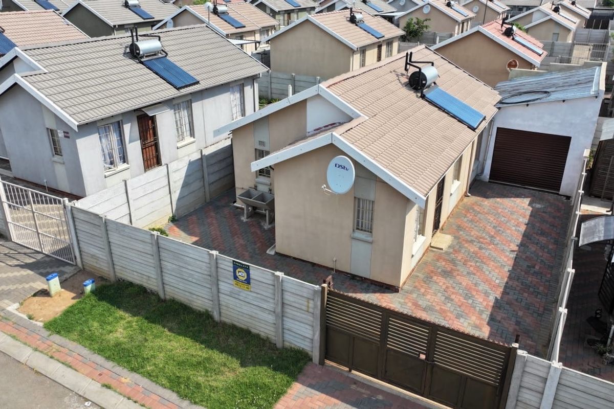 Aerial view of a residential property featuring a tiled roof with solar water heater, paved driveway, secure boundary walling, and a private garage.