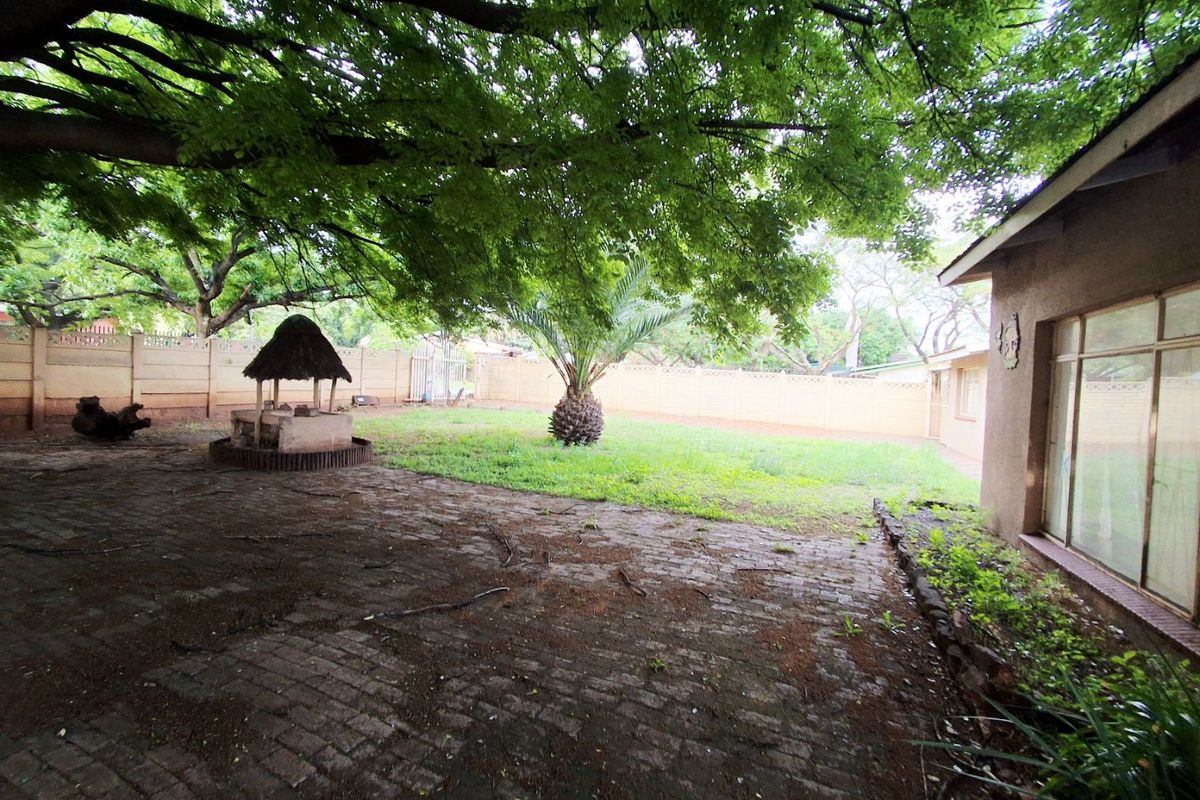 Spacious rear garden featuring a paved patio area, a thatched lapa, and mature trees enclosed by a precast concrete boundary wall.