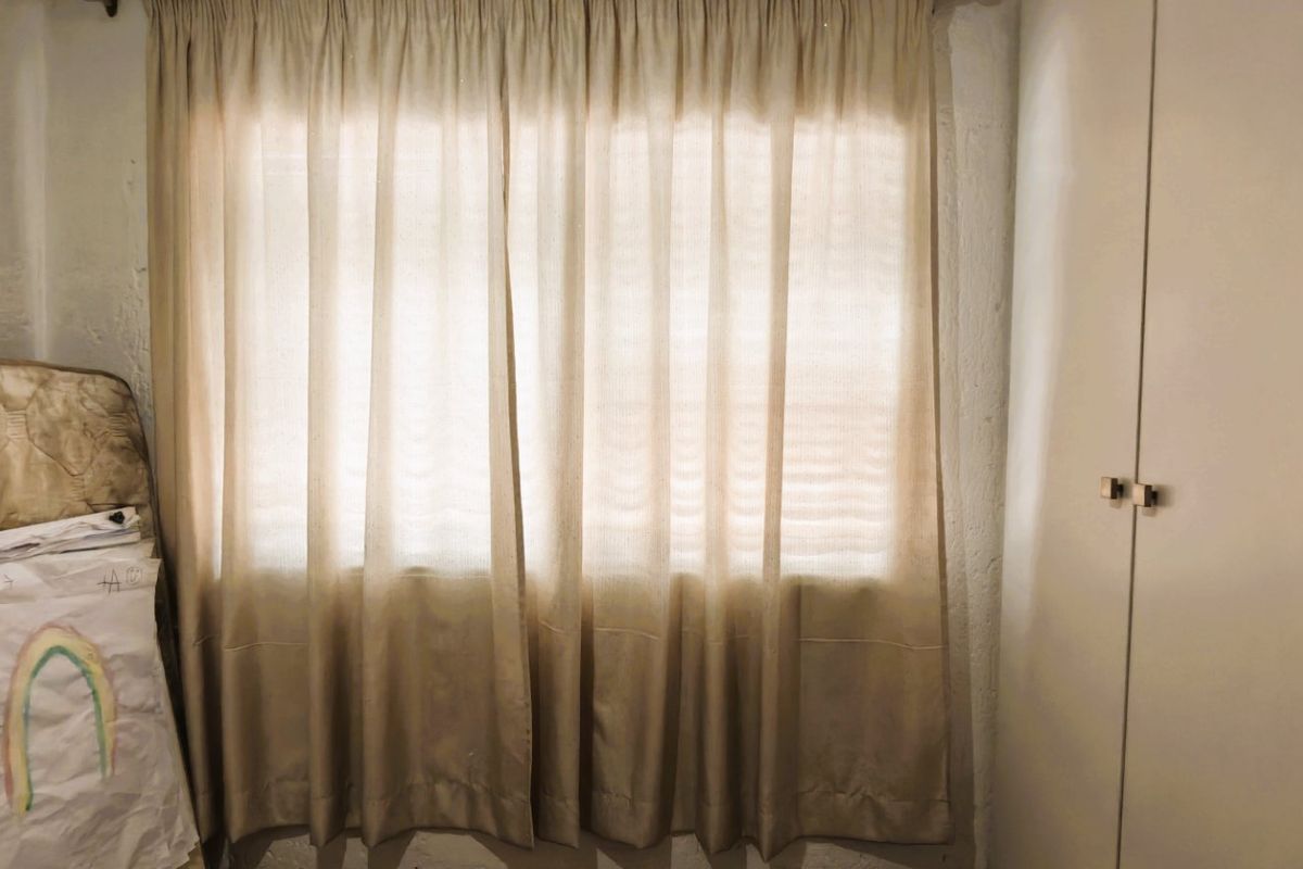 A bedroom featuring a built-in wardrobe and window with curtains, showing signs of wear on the wall surfaces.