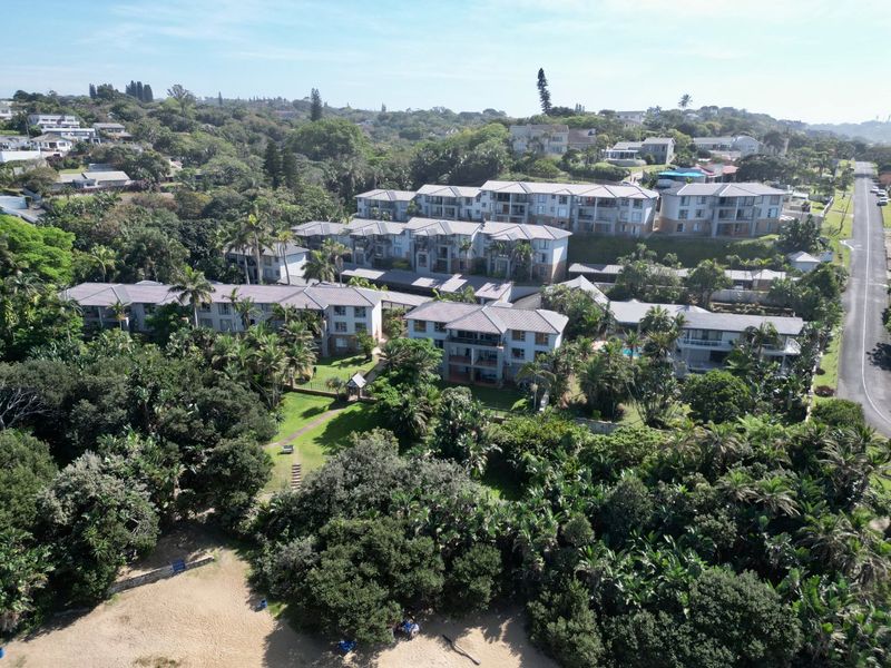 Aerial view of a residential apartment complex set within lush, landscaped grounds, featuring multiple buildings with tiled roofs and proximity to coastal vegetation.