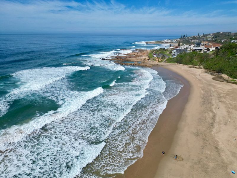 An aerial view of the coastline, showcasing the sandy beach, ocean waves, and surrounding coastal residential properties.