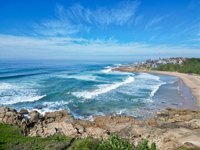A scenic coastal view overlooking the ocean, rocky shoreline, and sandy beach with residential properties visible in the distance.