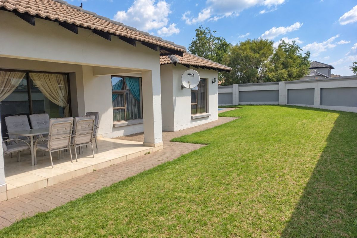 Rear view of the property featuring a covered patio, paved walkway, well-maintained lawn, and secure boundary wall.