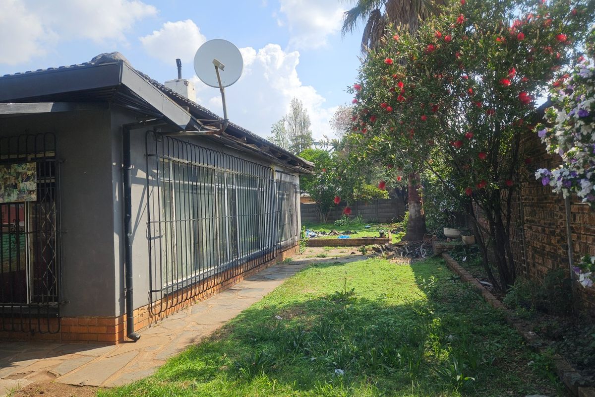 Rear view of the property featuring a paved walkway, security bars on windows, satellite dish, and a garden area with established trees and shrubs.